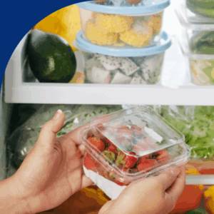 Hands holding strawberries in front of a refrigerator filled with fresh fruits and vegetables.